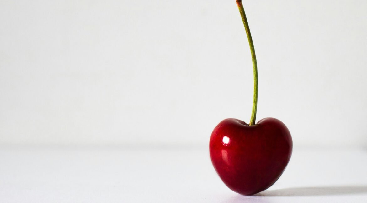 Cereja vermelha em formato de coração, com um pé comprido, destacando-se sobre um fundo branco. A fruta apresenta uma cor intensa e brilhante, simbolizando vitalidade e energia.