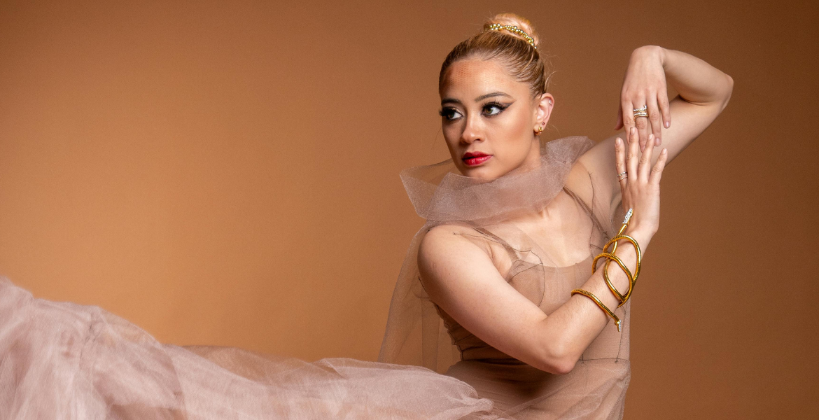 Mulher em pose de dança contemporânea, vestindo um vestido volumoso cor de nude, com os braços elevados e expressão de leveza.
